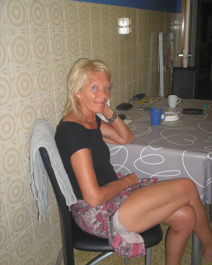 Femme aux cheveux blonds platine, portant un haut noir et une jupe noire, assise sur une chaise dans une cuisine, avec une table devant elle ayant des tasses et des objets divers.