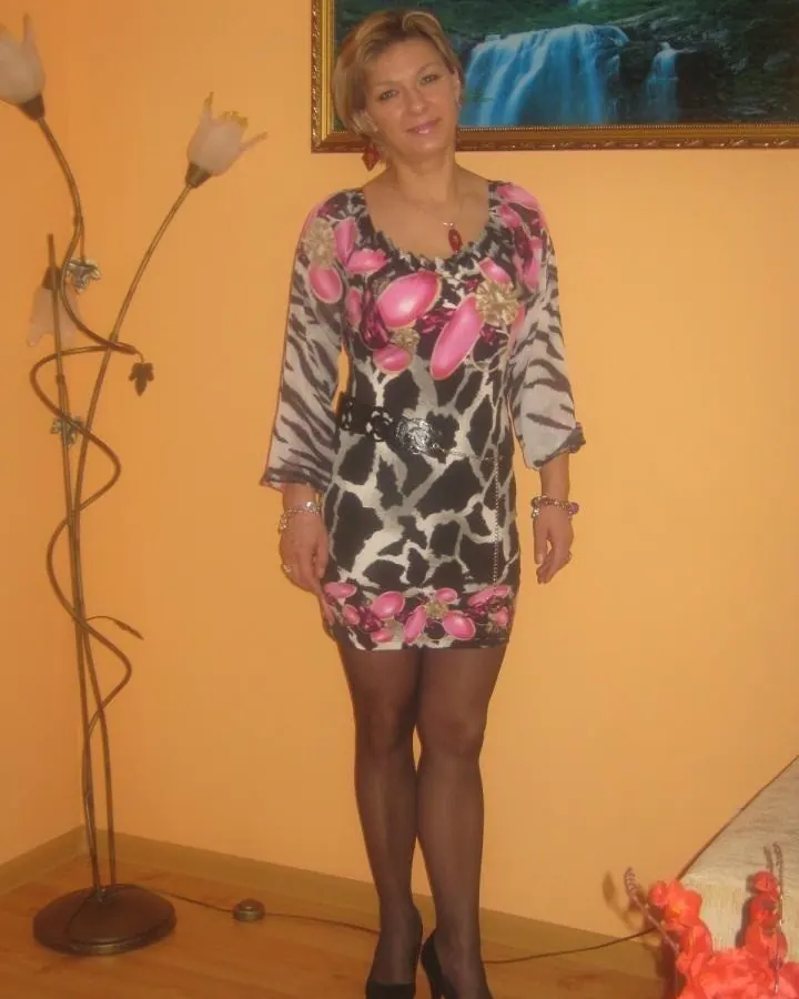 Femme blonde en robe imprimée léopard et fleurs roses, collants noirs et escarpins, debout dans une pièce aux murs orange avec une lampe décorative et un tableau de cascade.