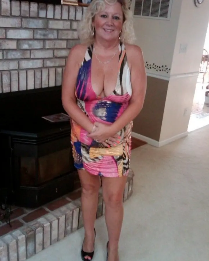 Femme blonde, en robe colorée, souriante, debout devant une cheminée en briques.