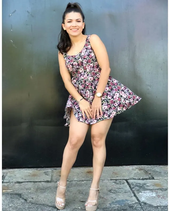 Femme aux cheveux châtains, en robe fleurie, souriante, debout contre un mur noir.