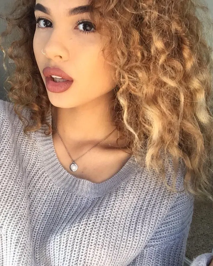 Femme aux cheveux blonds en tenue bleue, debout devant une grille, tirant ses cheveux, regardant l'appareil.