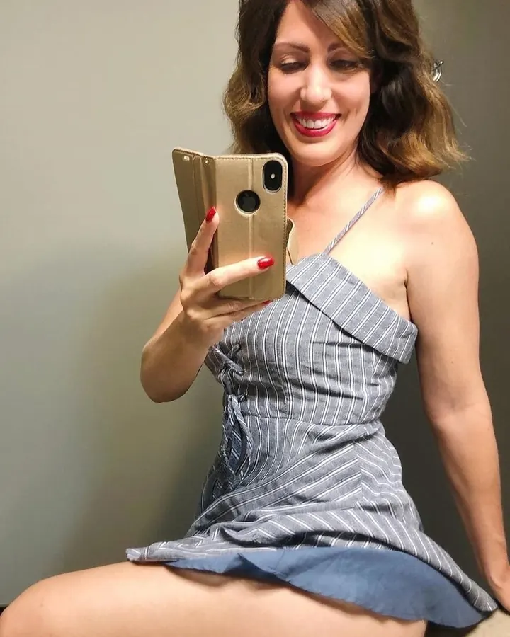 Femme aux cheveux châtains en robe grise, assise, prenant un selfie avec un téléphone doré, souriante.
