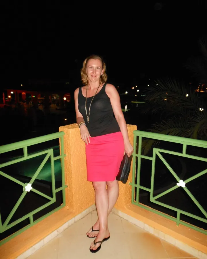 Femme aux cheveux blonds portant un haut noir et une jupe rose, debout sur une terrasse la nuit, souriante.