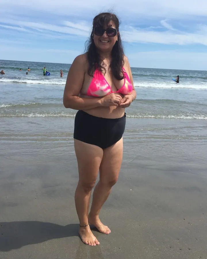 Femme blonde en bikini rose et noir, souriante, debout sur une plage.