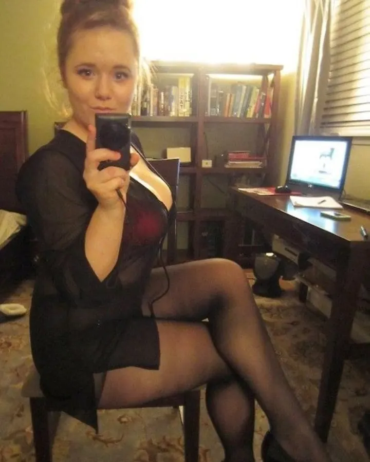 Jeune femme en robe noire transparente et collants, assise sur une chaise, prenant un selfie dans une chambre avec une bibliothèque en arrière-plan.