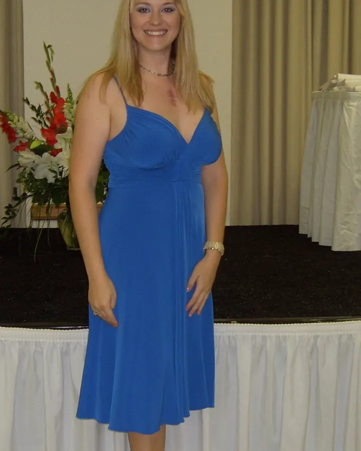 Femme blonde en robe bleue, souriante, debout devant une table décorée de fleurs.