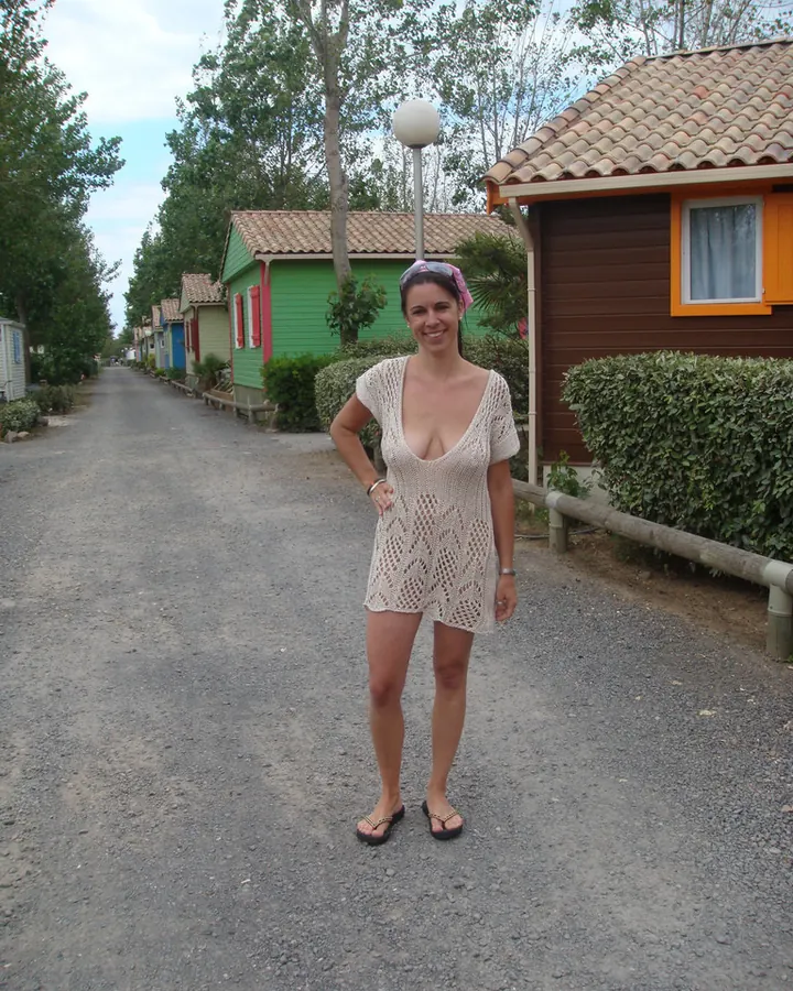 Femme souriante en robe tricotée claire, debout sur un chemin de campagne entre des chalets colorés.