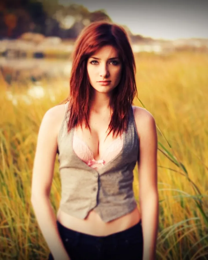 Femme aux cheveux roux, en gilet gris ouvert sur un soutien-gorge rose, debout dans un champ de blé doré, regardant directement l'appareil.