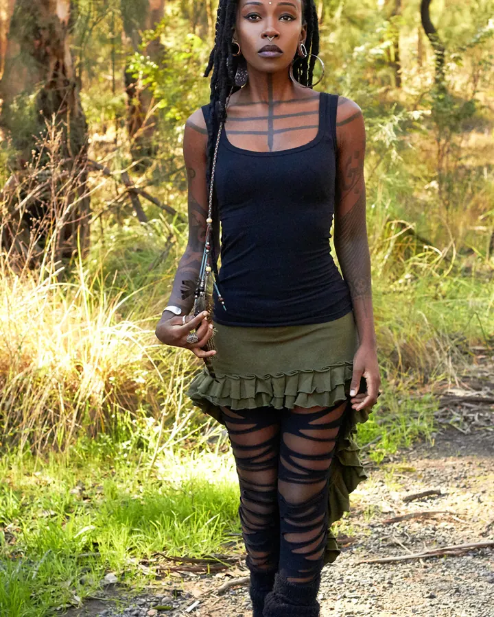 Femme aux cheveux noirs en dreadlocks, en haut noir et jupe verte à volants, pantalon en maille noire, posant dans une forêt.