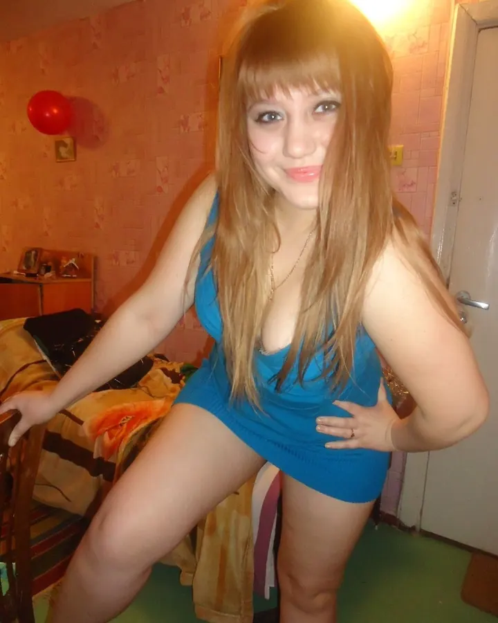 Femme aux cheveux blonds, en mini-robe bleue ajustée, posant dans une chambre avec un ballon rouge en arrière-plan.