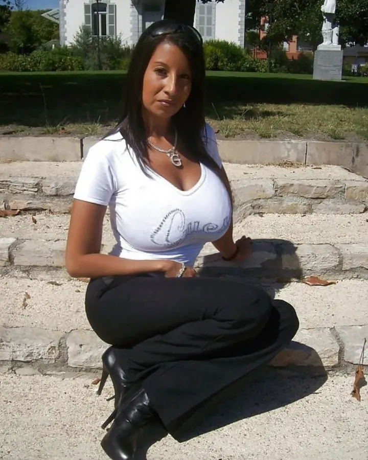 Femme aux cheveux bruns noirs, en T-shirt blanc à col V, en pantalon noir, assise sur des marches en pierre, souriant à l'appareil.