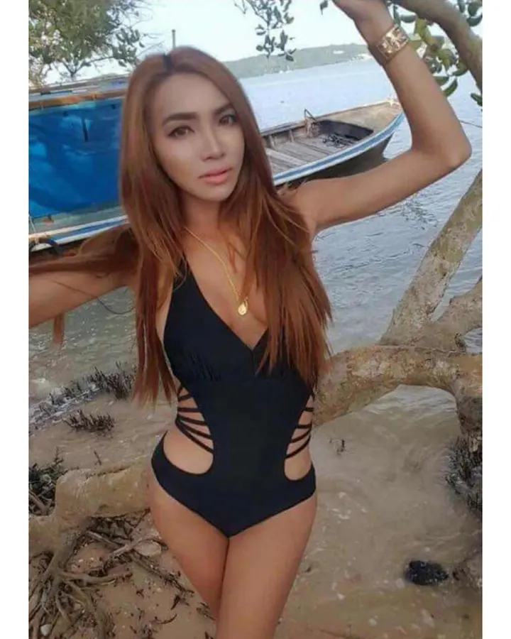 Femme aux cheveux bruns châtains, en maillot de bain noir avec découpes latérales, posant sur une plage, une main sur une branche d'arbre, un bateau en arrière-plan.