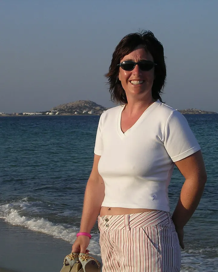 Femme aux cheveux bruns, vêtue d'un haut blanc, posant sur une plage avec des lunettes de soleil.