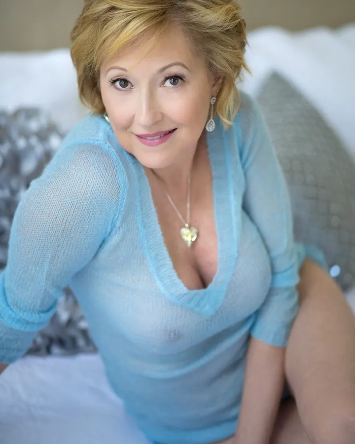 Femme aux cheveux blonds, vêtue d'un pull bleu clair, assise sur un lit avec un coussin gris.