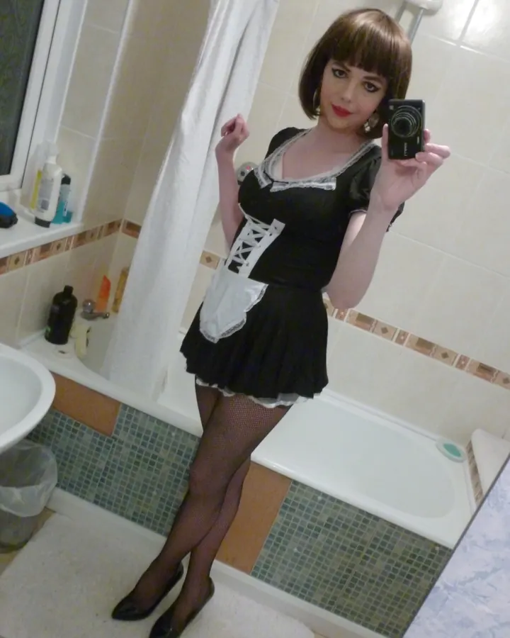 Femme aux cheveux bruns, vêtue d'une robe noire à lacets, tenant un appareil photo dans une salle de bain.