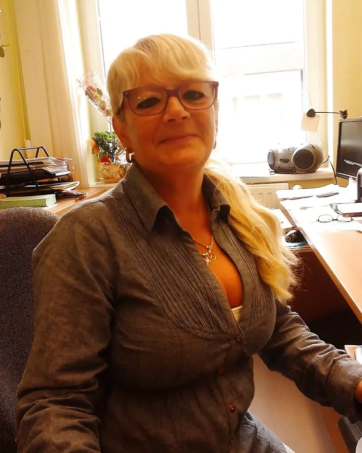 Femme blonde en chemise grise, lunettes, assise devant un bureau, regard direct