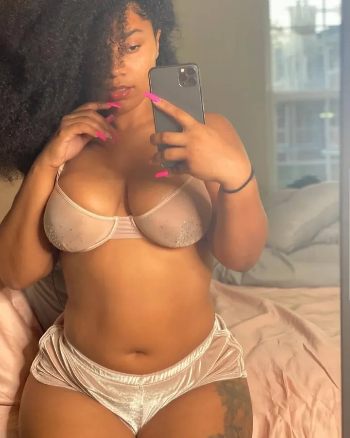 Femme en lingerie rose pâle, prenant un selfie, lit en arrière-plan, regard direct