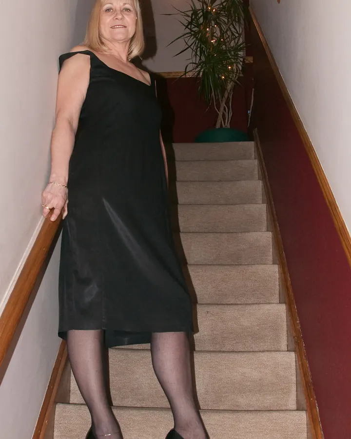 Femme aux cheveux blonds en robe noire, descendant un escalier, regardant l'objectif.