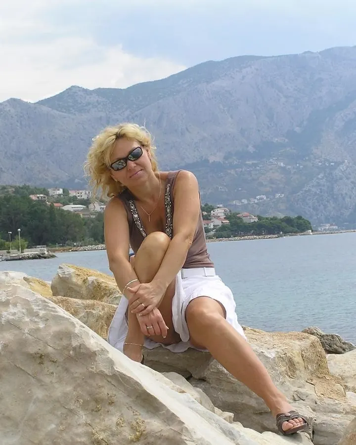 Femme aux cheveux blonds platine en haut marron, assise sur des rochers près d'une mer, portant des lunettes de soleil.