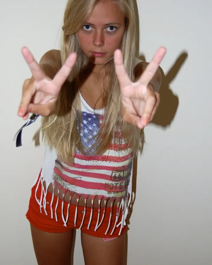 Jeune femme blonde en tenue décontractée avec drapeau américain, faisant le signe de la victoire avec les deux mains