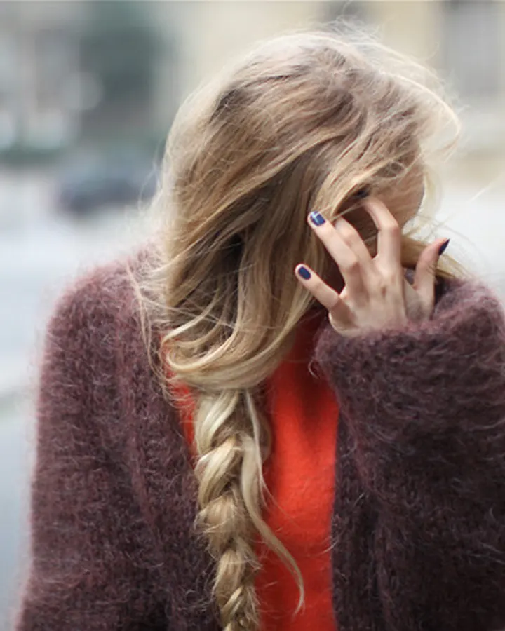 Femme blonde aux cheveux longs, vêtue d'un pull orange sous une veste marron, se tenant de profil avec une main devant le visage, sur une rue extérieure.