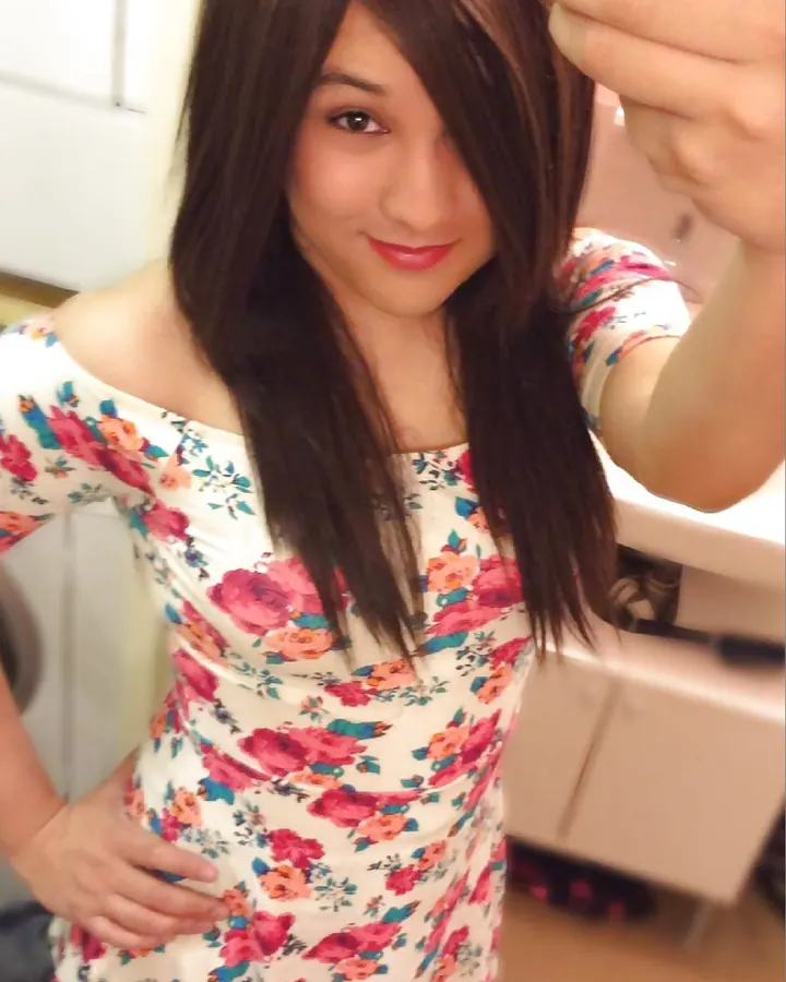 Femme aux cheveux châtains portant une robe fleurie blanche avec des roses rouges et oranges, prenant un selfie devant un miroir.