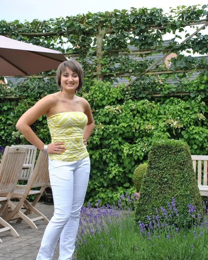 Jeune femme aux cheveux châtains en haut noir et short léopard, debout contre un mur blanc, regardant l'appareil avec une posture séduisante.
