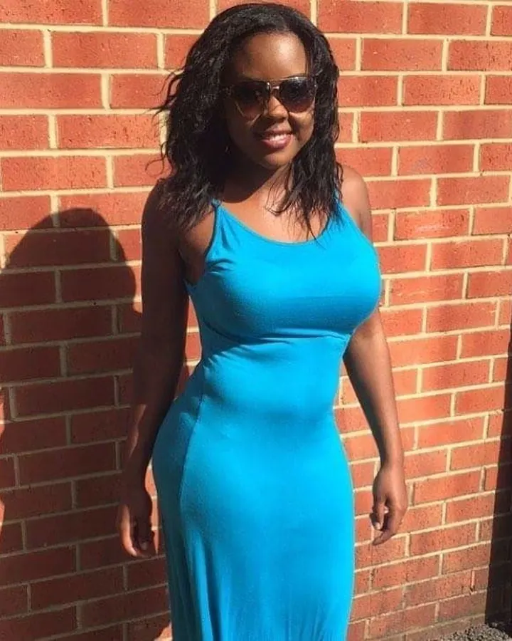 Femme noire en robe bleue ajustée, souriante, devant un mur de briques rouges, portant des lunettes de soleil.
