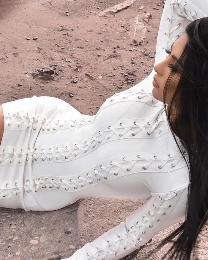 Femme aux longs cheveux noirs, allongée sur un sol terreux, portant une combinaison blanche à lacets, regardant vers le côté.