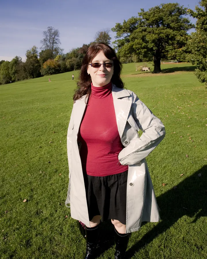 Femme en robe rouge à col roulé, portant un trench beige, debout dans un parc verdoyant, souriante, sous un ciel bleu.