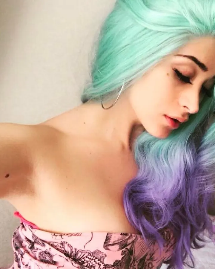 Jeune femme aux cheveux noirs, vue de profil, avec une perruque turquoise et violette, portant un haut rose imprimé.
