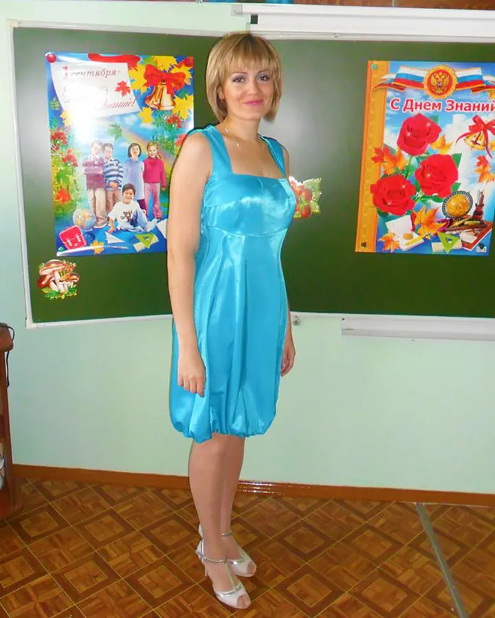 Femme aux cheveux blonds courts, souriante, portant une robe bleue satinée, debout devant un tableau noir décoré pour la rentrée scolaire.
