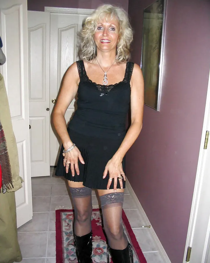 Femme aux cheveux blonds, portant une robe noire avec dentelle, debout dans un couloir intérieur.